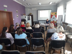 Na zdjęciu widać osoby siedzące na krzesłach, a tle prezentację multimedialną,.