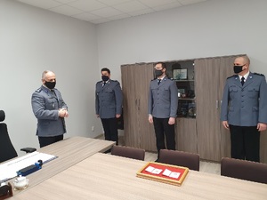 Na zdjęciu widać czterech umundurowanych wyjściowo policjantów w pomieszczeniu. Trzech z nich stoi  w szeregu, czwarty stoi z boku zwrócony do pozostałej trójki.