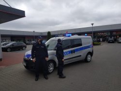 Zdjęcie przedstawia dwóch umundurowanych policjantów stojących przed oznakowanym radiowozem, w tle widac parking i sklepy.