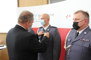 Na zdjęciu widać dwóch policjantów stojących przodem oraz osobę, która wręcza im odznaczenia.