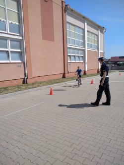 Na zdjęciu widoczny jest policjant ruchu drogowego egzaminujący na kartę rowerową. Na dalszym planie widać dziecko jadące na rowerze między pachołkami.